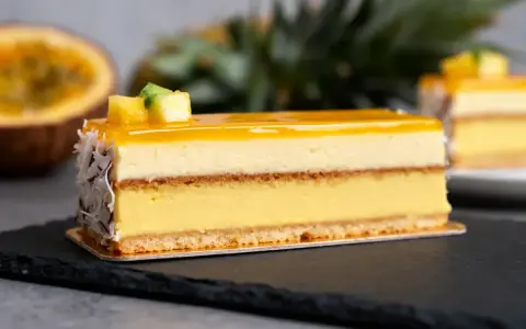 Un délice tropical : un entremets aux couches crémeuses, surmonté d'un glacage miroir jaune éclatant, décoré de fruits et de noix de coco râpées.