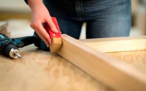 Eine Hand schleift mit einem roten Schleifklotz ein Holzstück. Daneben liegt ein Akkuschrauber.