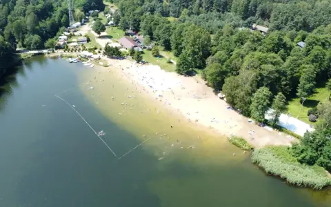 Słoneczny dzień nad jeziorem Rogoźnik. Ludzie kąpią się i opalają na piaszczystej plaży, otoczonej zielonym lasem.