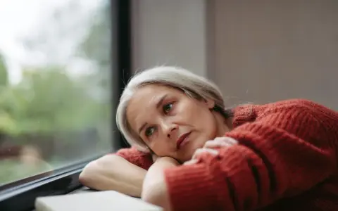 Donna con capelli grigi guarda fuori dalla finestra, pensierosa. Potrebbe cercare rimedi per la depressione estiva.