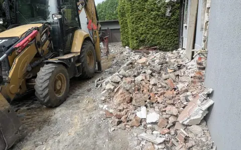 Żółta koparko-ładowarka pracuje przy wyburzeniu domu. Gruz i cegły leżą na ziemi. Koszt wyburzenia domu jest wysoki.