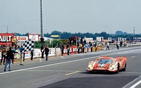 Ein roter Porsche Rennwagen mit der Startnummer 23 überquert die Ziellinie, während eine karierte Flagge geschwenkt wird.