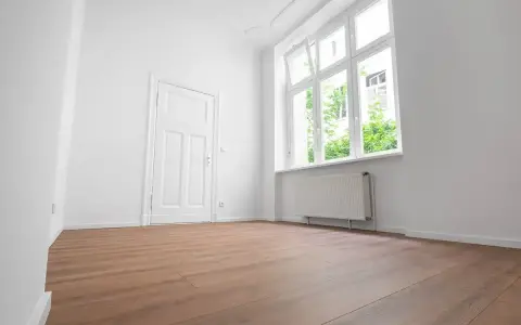 Puste, jasne pomieszczenie z białymi ścianami, drzwiami i oknem. Drewniana podłoga i grzejnik sugerują, że to gotowe do zamieszkania mieszkanie socjalne.