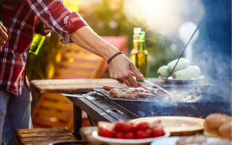 Hombre asa salchichas envueltas en tocino, ideal para saber qué llevar a una barbacoa para sorprender. Hay cervezas y tomates en la mesa.