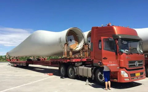 Pomarańczowy tir przewozi gigantyczne łopaty turbin wiatrowych. Kierowca przygotowuje się do dalszej podróży.