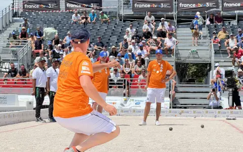Joueur de pétanque concentré, cherchant à faire le vide dans sa tête avant de lancer la boule. Le public est installé dans les gradins derrière lui.