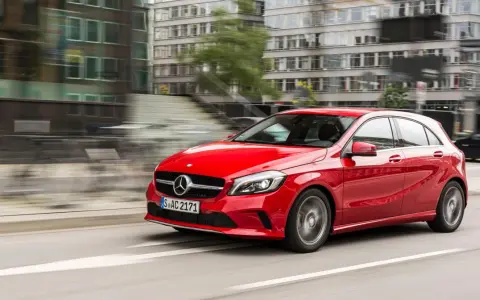 Czerwony Mercedes-Benz W176 pędzi ulicą miasta, mijając nowoczesne budynki.