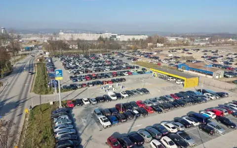 Duży parking przy krakow katowice airport. Wiele samochodów zaparkowanych w rzędach, w tle budynki lotniska.
