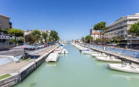 La darsena di Riccione, con barche ormeggiate lungo il canale, palazzi e alberi verdi sotto un cielo azzurro.