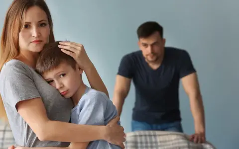 Smutna kobieta tuli syna, podczas gdy partner patrzy z dystansu. Trudno jej zaakceptować dziecko partnera.