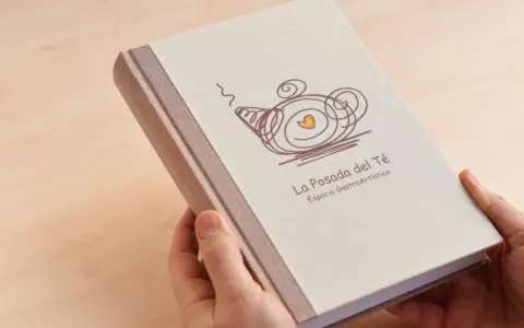 Manos sostienen un libro con tapa de tela, con un diseño de tetera y el texto "La Posada del Té". Una idea para como encuadernar un trabajo de forma original.