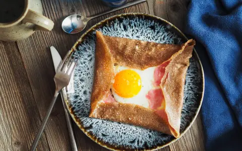 Une galette de sarrasin garnie d'un œuf au plat et de jambon, un des nombreux types de crêpes salées. Servi sur une assiette avec des couverts.