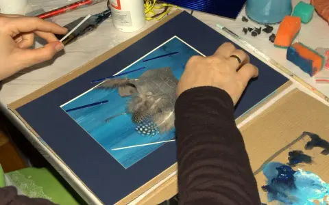 Ręce tworzą artystyczny asamblaż z piór i niebieskich elementów na tle malowanym na niebiesko.