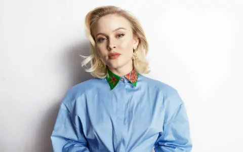 Zara Larsson, känd för sin musik, bär en blå skjorta med en färgglad krage. Hennes skivbolag har säkert hjälpt henne nå framgång.