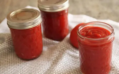 Trois bocaux de ketchup maison, un rouge vif, reposent sur une serviette à motifs. Un tomate entière est visible.