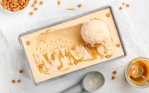 Ein Blech mit cremigem, hellbraunem Salted Caramel Eis, garniert mit Karamellsauce und kleinen Karamellwürfeln. Eine Eiskugel liegt darauf.