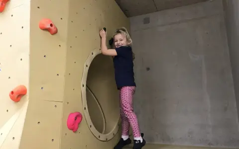 Ein fröhliches Mädchen klettert in einer Kletterhalle für Kinder. Sie lächelt und hält sich an einem Griff fest.