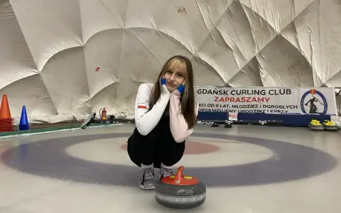 Młoda sportsmenka na lodzie, gotowa do gry w curling. W tle baner "Gdańsk Curling Club Zapraszamy".