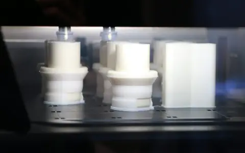 Białe obiekty w drukarce 3D, z widocznym elementem przypominającym 3d print raft, tworzą się warstwa po warstwie.