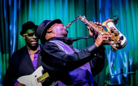 Muzyk gra na saksofonie, tworząc nowoczesny jazz. W tle widać gitarzystę.