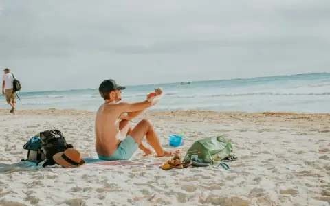Mężczyzna robi zdjęcie telefonem na plaży, marząc o najkrótszych lotach z Polski na wakacje. Obok leżą torba, kapelusz i sandały.