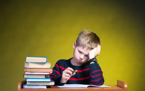 Zmęczony chłopiec z podpartą głową przy biurku, gdy szkoła nie realizuje zaleceń poradni. Stos książek obok.