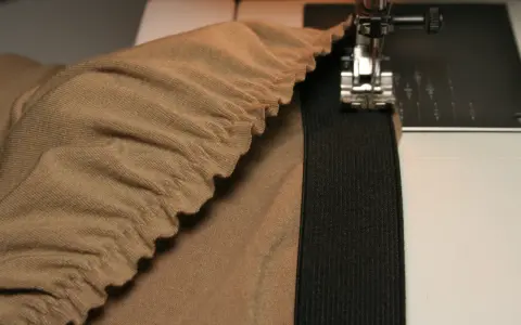 Machine à coudre en train de coudre un élastique sur un tissu beige froncé. Le pied presseur est visible, ainsi que l'aiguille.