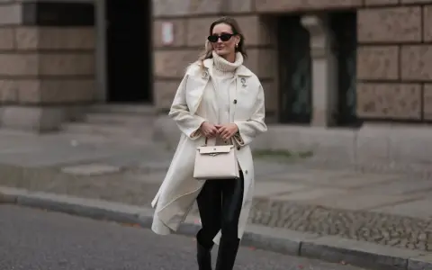 Elegancka kobieta w kremowym płaszczu, golfie i skórzanych legginsach, z beżową torebką. Stylowe skórzane legginsy stylizacje.