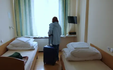 Kobieta z walizką przy oknie, gotowa by dowiedzieć się, jak wyjechać do sanatorium. Pokój z dwoma łóżkami.