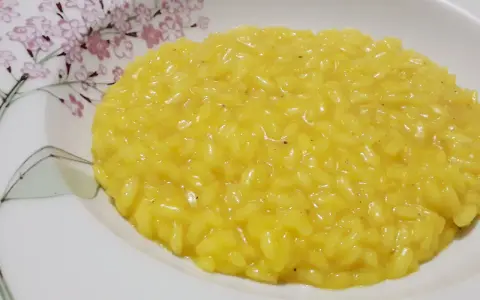 Un cremoso risotto allo zafferano, servito in un piatto bianco decorato con fiori rosa e rami verdi.