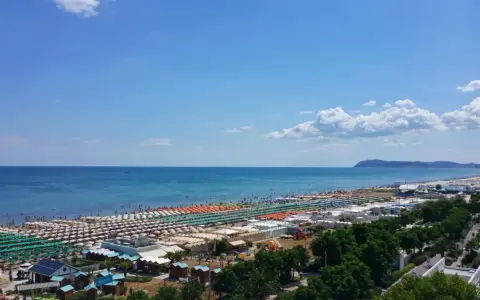 Ampia spiaggia di Riccione con file ordinate di ombrelloni e lettini, bagnanti che si godono il sole e l'acqua cristallina del mare Adriatico sotto un cielo azzurro.