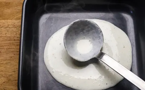 Eine Schöpfkelle verteilt cremige Bechamel-Sauce in einer schwarzen Auflaufform.
