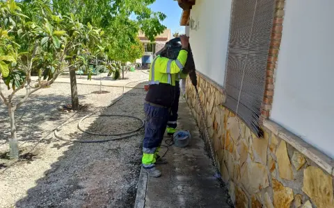 Trabajador con equipo de protección realiza trabajos de inyección de cemento en la base de una pared exterior, rodeado de vegetación.