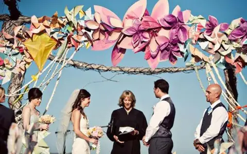 Boda al aire libre con un arco decorado con manualidades de papel coloridas. La pareja se da el sí ante un oficiante.