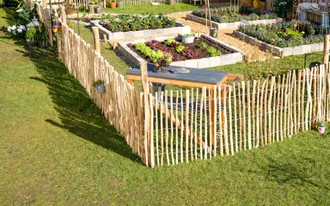 Ein rustikaler Staketenzaun wird befestigt, um einen liebevoll angelegten Gemüsegarten mit Hochbeeten zu umzäunen.