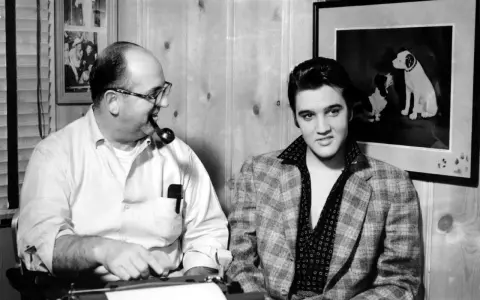 Elvis Presley und Colonel Parker, sein Manager, sitzen nebeneinander. Parker raucht Pfeife, Elvis blickt in die Kamera.