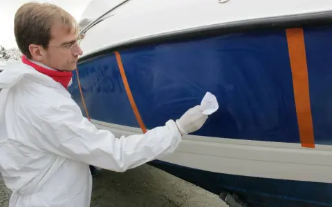 Un homme en combinaison blanche effectue la mise en cire d'un bateau bleu, protégeant la coque pour l'hiver.
