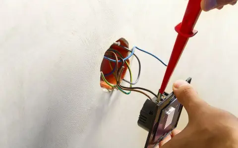 Ręce montują włącznik światła, podłączając przewody. Widać instalację elektryczną i narzędzie.