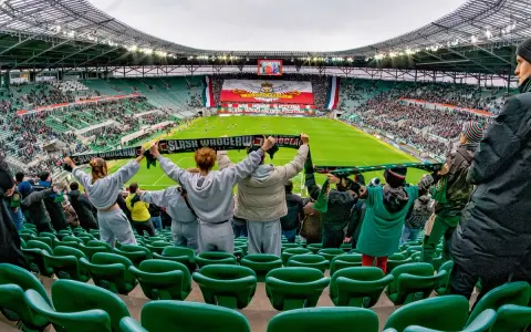 Gwardia Opole kibicuje na stadionie, trzymając szaliki. Na boisku trwa mecz, a trybuny wypełniają fani.