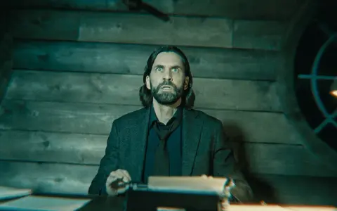Alan Wake 2: un hombre con barba y pelo largo, con expresión de sorpresa, sentado frente a una máquina de escribir en una habitación de madera.