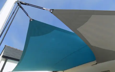 Comment installer un voile d'ombrage sur terrasse : deux voiles, bleu et gris, fixés par des cordages et des poulies à un mât et à la maison.