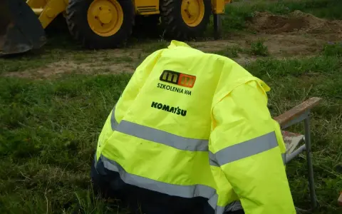 Żółta kurtka z logo "SZKOLENIA MM KOMATSU" leży na krześle. W tle koparko-ładowarka. Idealna na pytania na koparko ładowarkę.
