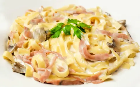 Tagliatelle z szynką i pieczarkami w kremowym sosie, ozdobione natką pietruszki. Pyszny makaron z mozzarellą w roli głównej.