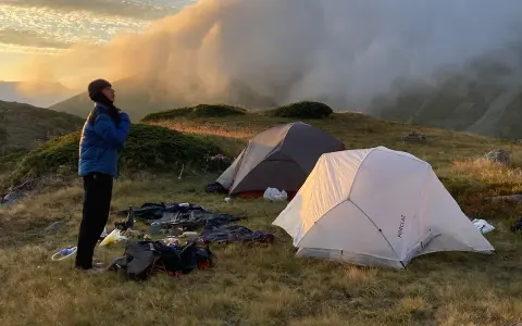 Une personne se tient près de tentes dans une aire de bivouac, profitant du lever du soleil sur les montagnes.