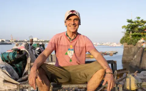 Le chanteur Mano Negra, souriant, assis sur un banc rouillé, avec un collier coloré et un t-shirt à motif poisson.
