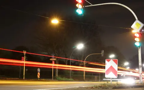 Nachts an einer Kreuzung mit roten Lichtspuren von Autos. Ampeln zeigen Grün und Rot. Ein Vorfahrtsschild und ein Richtungspfeil sind zu sehen. 132 bkat.