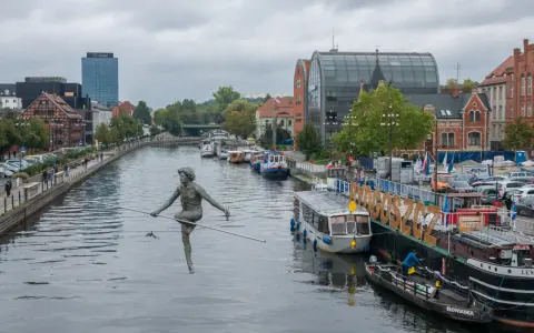 Bydgoszcz atrakcje: rzeźba na linie nad Brdą, cumujące statki i budynki nad rzeką.