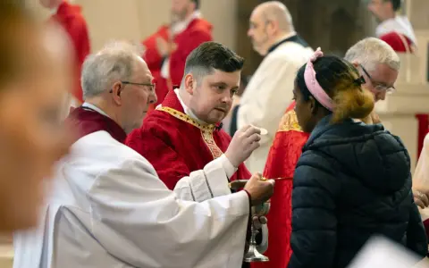 Clergy in red robes perform communion, a sacred act that can be seen as a form of evangelizing, sharing faith with a young woman.