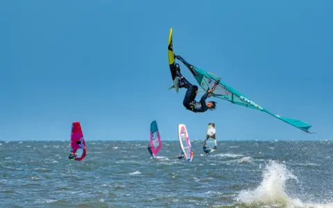 Windsurfer vollführt spektakulären Sprung über den Wellen. Mehrere andere Windsurfer sind auf dem Wasser zu sehen, was auf beliebte Windsurfing Spots hindeutet.