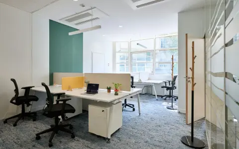 Un bureau chaleureux avec des postes de travail modernes, des cloisons acoustiques et un tapis bleu doux. L'espace est lumineux grâce aux grandes fenêtres.
